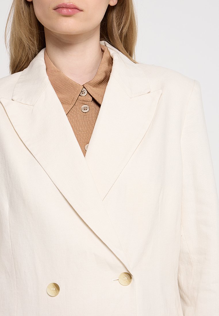 Blazer double boutonné de couleur crème en tissu texturé, avec des revers proéminents et deux grands boutons beiges. Une chemise à col marron clair est visible en dessous.
