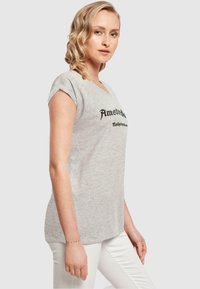 Tricou din bumbac gri cu decolteu în V și mâneci răsucite, având text negru care spune „Amsterdam Olanda”. Textură netedă, croială lejeră.
