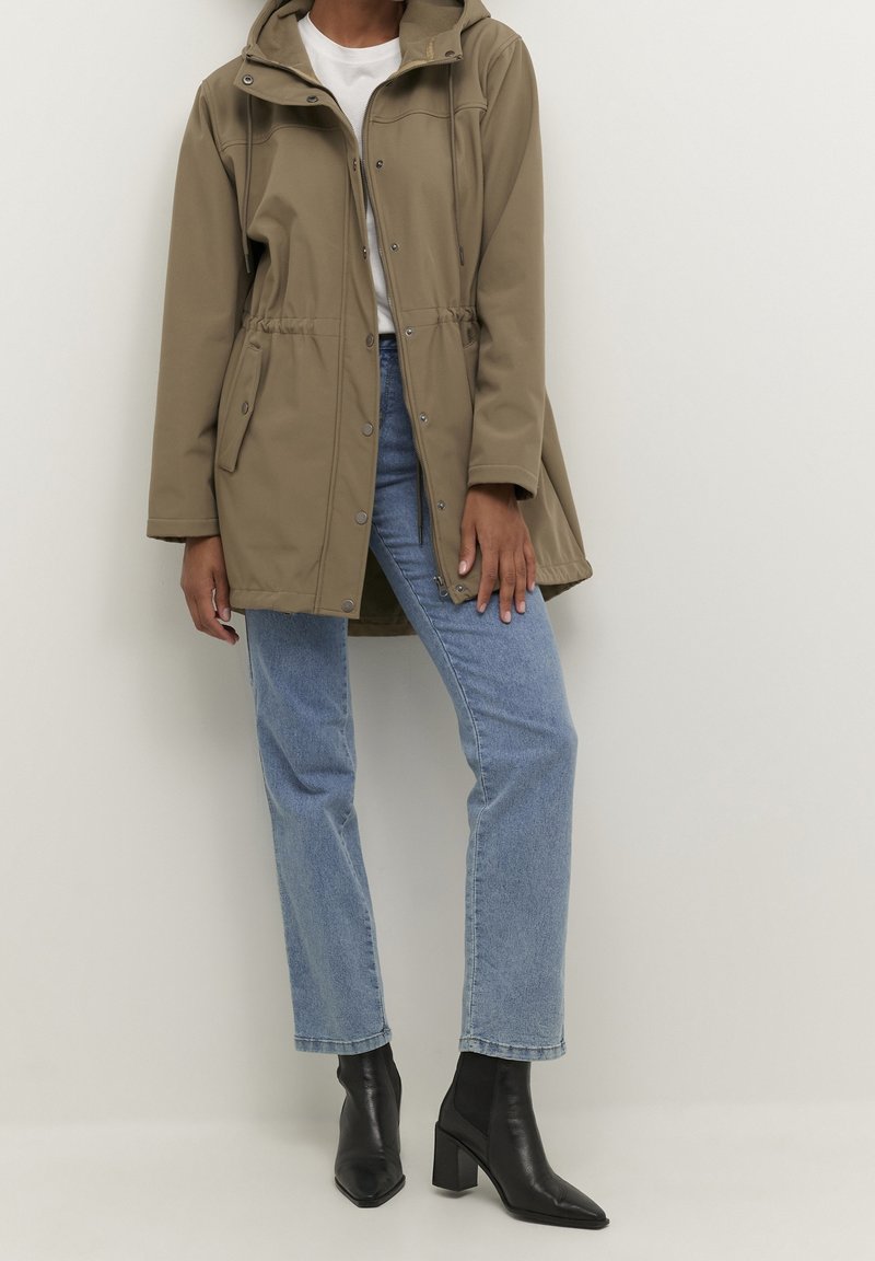 Giacca impermeabile color khaki con cappuccio, vita elastica, bottoni a pressione e tasche laterali, indossata sopra jeans azzurri e stivali neri con tacco.