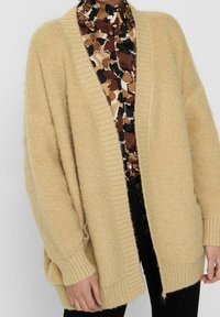 Béžový, nadměrné velikosti kardigan s texturovaným úpletem, otevřený vpředu a s žebrovanými manžetami. Nošený přes vzorovanou blůzu, s uvolněným střihem a spadlými rameny.
