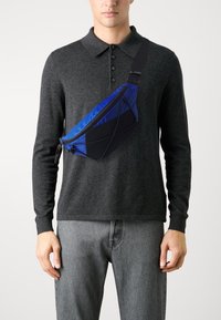 Schwarze und blaue Umhängetasche aus Nylon mit einem Reißverschlussfach, dreieckiger Form und verstellbarem Tragegurt, die über einem grauen Pullover getragen wird.