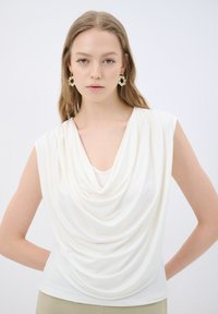 Cowl-Neck-Top aus cremefarbenem Stoff mit leichter Drapierung, ärmellosem Schnitt und weicher Textur. Klarer Hintergrund, um den Fokus auf das Kleidungsstück zu legen.