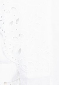 Tissu blanc présentant une broderie complexe avec des motifs découpés. Texture douce avec un léger éclat, détails le long du bord.