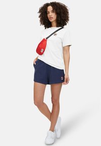 Witte katoenen t-shirt met een klein logo, gecombineerd met marineblauwe shorts en witte sneakers. Een rode schoudertas wordt over één schouder gedragen.