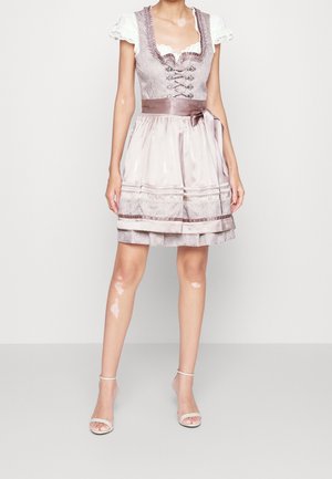 Kobieta ubrana w jasnofioletową tradycyjną sukienkę dirndl z gorsetem sznurowanym na przodzie, białymi bufkami na rękawach, satynowym fartuchem oraz białymi butami na obcasie z paskiem wokół kostki.