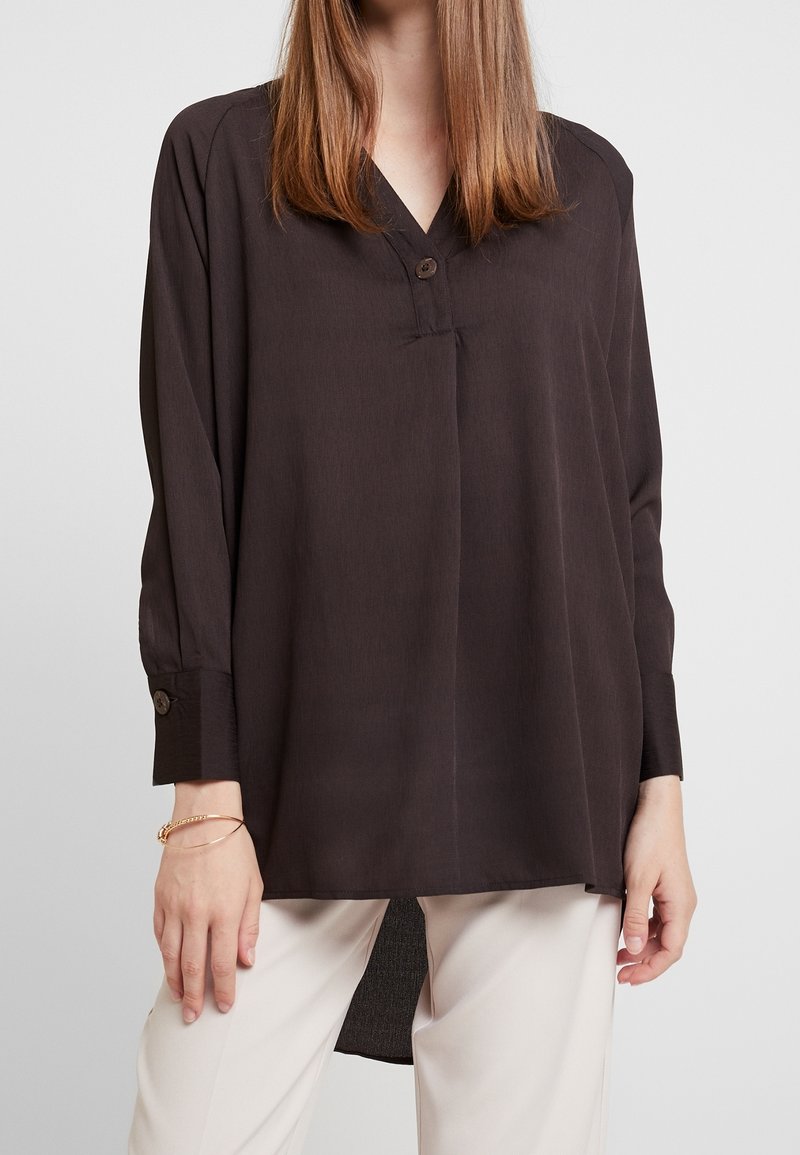 Femme portant un chemisier noir ample à manches longues avec un col boutonné et un pantalon blanc sur un fond uni.