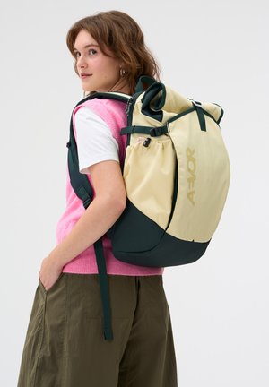 Junge Frau trägt eine rosa Weste und olivgrüne Hose und hat einen beigen und dunkelgrünen AEOR-Rucksack über der Schulter vor weißem Hintergrund.