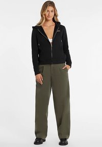 Felpa nera con zip e coulisse regolabili, tasche frontali e polsini a costine, abbinata a pantaloni larghi verde oliva e stivali neri.