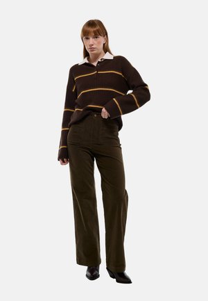 Pull en beige rayé marron avec col crème, associé à un pantalon en velours côtelé olive. Le mannequin pose avec confiance, portant des bottes à talons noires.