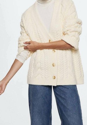Persoon draagt een crèmekleurige kabelgebreide vest met knopen over een witte coltrui, gecombineerd met loszittende blauwe spijkerbroek.