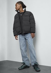 Zwarte pufferjack met elastische manchetten, gecombineerd met een zwart shirt en lichtblauwe jeans. Draagt groene sneakers met zwarte accenten.