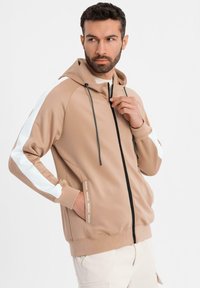 Beige zip-up hoodie med dragsko i huvan, vita sidosträck, och ribbade muddar. Har en mjuk, lättviktig tygkvalitet och logotypdetalj.