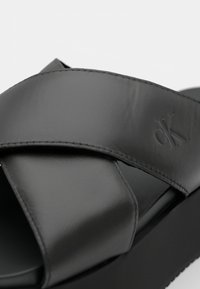 Černá pantofle na podpatku s lesklým koženým svrchním dílem, který má překrývající se křížový páskový design. Podešev je tlustá a texturovaná.