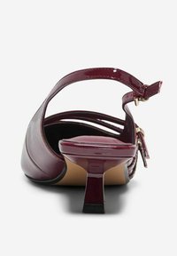 Sandale à talon haut en cuir verni bourgogne avec un talon fin et une sangle de cheville réglable avec des accessoires en métal doré. Fini lisse et brillant.