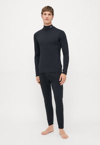 Homme debout pieds nus portant un haut à manches longues noir ajusté et un pantalon noir assorti, sur un fond blanc uni.