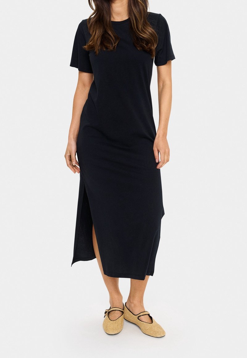 Robe noire à manches courtes, longueur midi, avec une fente latérale, en tissu doux. Portée avec des chaussures beige tissées sans lacets.