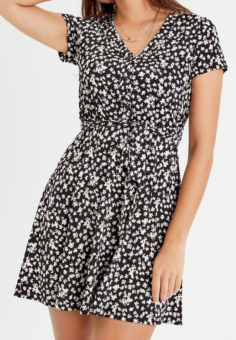 Femme portant une robe noire courte avec un imprimé floral blanc, à manches courtes, décolleté en V et ceinturée à la taille, accessoirisée avec des colliers en or superposés.