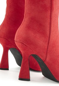 Red suede high-heeled boots with a textured finish, featuring a sleek silhouette and a curvy heel design. Black rubber soles.