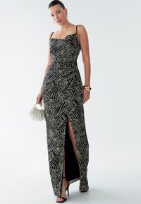 Vestido maxi en negro y crema con patrón de olas, cuenta con tirantes finos, una cintura ajustada y una abertura alta en el muslo. Los accesorios incluyen un bolso.