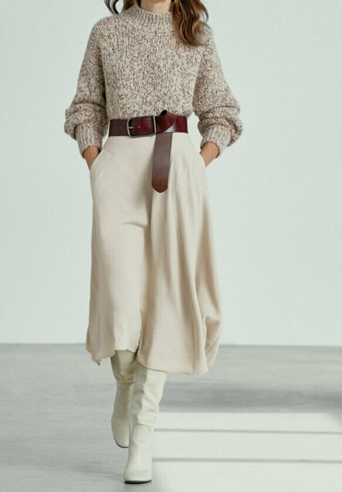 Beige gebreide trui met een hoge halslijn, gecombineerd met een vloeiende crèmekleurige rok die is getailleerd met een brede bruine riem. Tot de knie hoge witte laarzen.