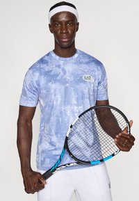 Ανοιχτό μπλε αθλητικό πουκάμισο με κοντά μανίκια, με υφή και αφηρημένο μοτίβο, και λογότυπο στο αριστερό στήθος. Λαβή ρακέτας τένις στο δεξί χέρι.