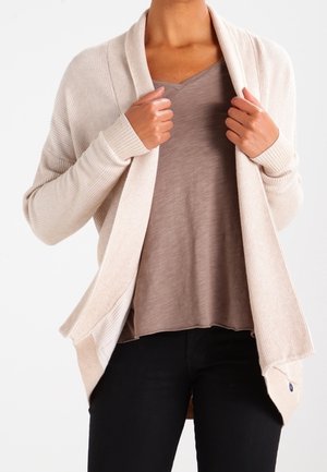 Cardigan côtelé beige avec un large devant ouvert, des manches longues et des poches latérales, porté sur un t-shirt blanc cassé à col en V et un pantalon noir.