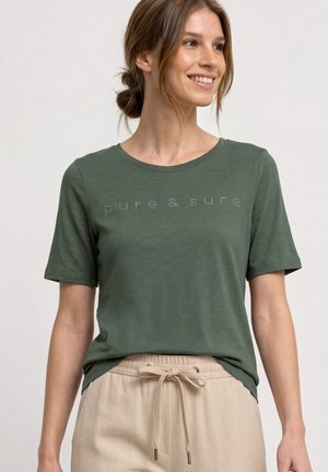 Kvinde smilende iført en grøn t-shirt med teksten "puro & sure" og beige snøre bukser mod en ensfarvet lys baggrund.