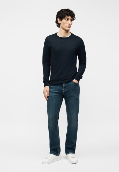 Άνδρας με σγουρά μαλλιά, φορώντας μαύρο πουλόβερ με μακριά μανίκια, μπλε τζιν, λευκά αθλητικά παπούτσια, στέκεται με το ένα χέρι στην τσέπη, μπροστά σε απλό φόντο.