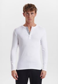 Hvid langærmet henley skjorte, som har knapper ved forkanten, en rund halsudskæring og et tekstureret sort stribedesign langs forkanten.