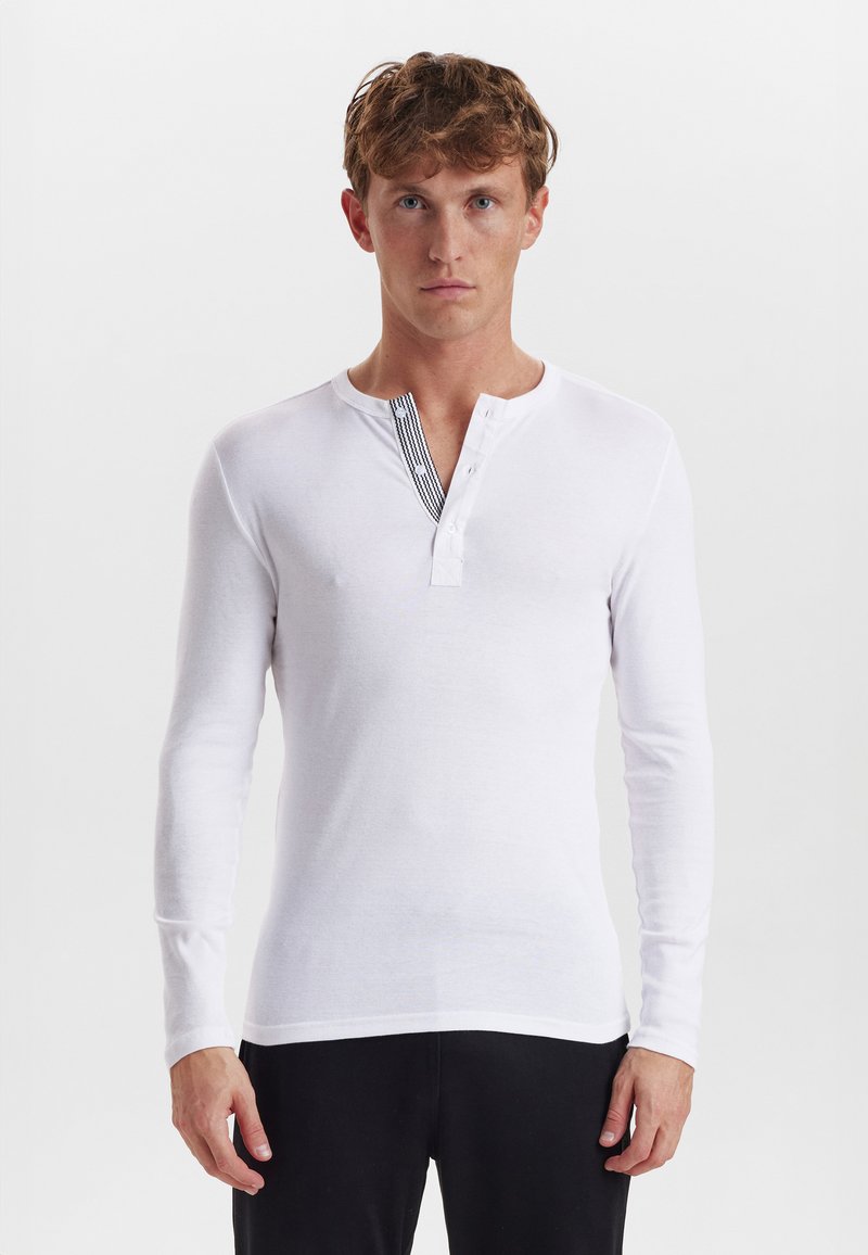 Hvid langærmet henley skjorte, som har knapper ved forkanten, en rund halsudskæring og et tekstureret sort stribedesign langs forkanten.