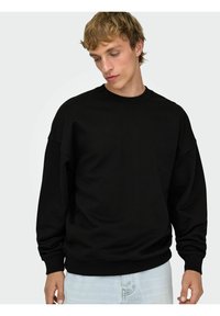 Felpa nera con scollo rotondo, spalle scese, polsini e orlo a costine. Tessuto morbido, vestibilità comoda. Indossata sopra jeans azzurri.