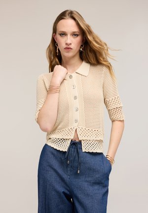 Junge Frau mit welligem Haar, die ein beiges Häkel-Top mit Knöpfen und blaue Hosen mit Kordelzug trägt, posiert mit der Hand nahe am Kinn vor neutralem Hintergrund.