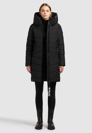 Schwarze Steppjacke mit Kapuze, mit gesteppter Optik, Frontreißverschluss und seitlichen Reißverschlusstaschen. Kombiniert mit schwarzen Leggings und Stiefeln.