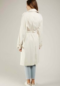 Trench-coat couleur crème avec une ceinture, présentant un design à double boutonnage, des poches à rabat et des poignets ajustables. Porté par-dessus un léger denim.