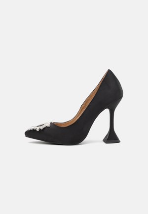 Chaussure à talon haut en satin noir avec un bout pointu, présentant un accent décoratif en forme de fleur argentée à l'avant et un design de talon triangulaire unique.