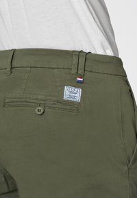 Primo piano di pantaloni verde oliva con un'etichetta USPA sulla tasca posteriore, che mette in risalto un bottone e parte di una camicia chiara sopra.
