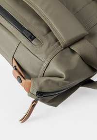 Zaino in nylon verde oliva con finitura testurizzata, dotato di una cerniera nera e dettagli in pelle beige, tra cui un bottone e un linguetta per il ritorno.