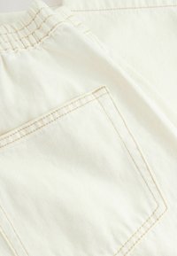 Gros plan sur un tissu en denim couleur crème montrant une taille élastique et une poche arrière avec des coutures beige.