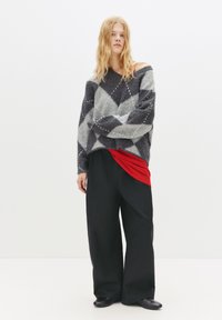 Pulover supradimensionat gri și negru din tricot cu model în diamant, purtat peste un top roșu și cu pantaloni largi negri. Textură fină, stil casual.