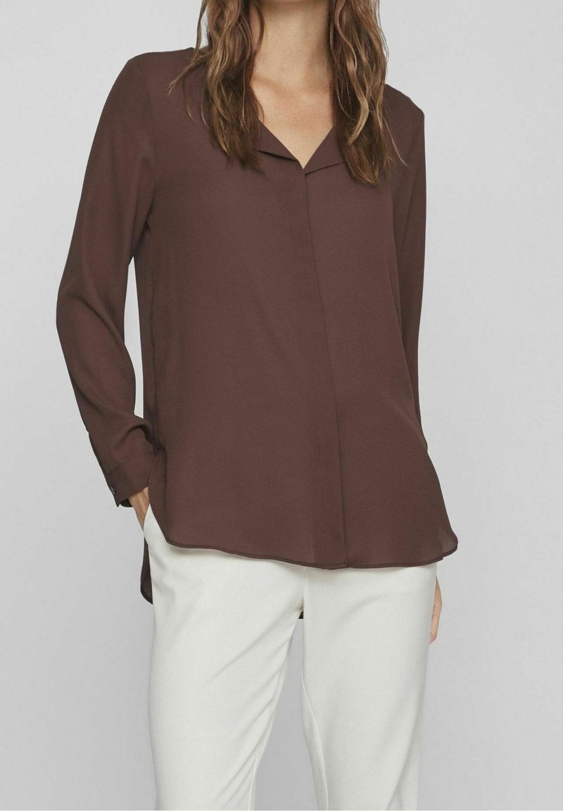 Brun blus med lång ärm, v-ringning och avslappnad passform. Tillverkad av lättviktigt tyg, har en rundad nederkant och en subtil glänsande textur.