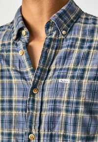 Camisa de cuadros abotonada en tonos azul, verde y amarillo. Material de algodón, cuello estándar, botones de madera y un parche de logo en el pecho.