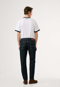 Biely polo-shirt s námorníckym lemovaním, v kombinácii s tmavomodrými džínami. Medzi charakteristiky patrí logo na zadnej strane a dve zadné vrecká so šitím.