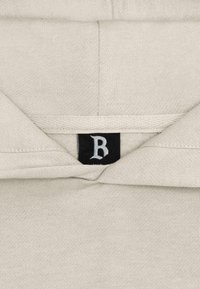 Hellbeige Kapuzenpullover mit einer weichen Textur, versehen mit einem schwarzen Etikett mit einem weißen Buchstaben "B" im Kragen, das ein minimalistisches Design zeigt.