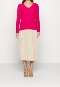 Světle růžový pletený svetr s V-výstřihem a dlouhými rukávy, doplněný o plisovanou béžovou midi sukni a světlé béžové sandály na řemínky.
