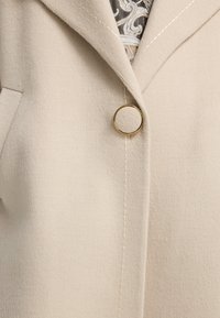 Lyse beige frakke lavet af blødt stof med klapper, der har en gylden, rund knap og synlige syningsdetaljer.