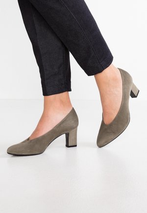 Füße in taupefarbenen Wildleder-Pumps mit niedrigem Absatz, kombiniert mit schwarzen, gekürzten Hosen vor weißem Hintergrund.