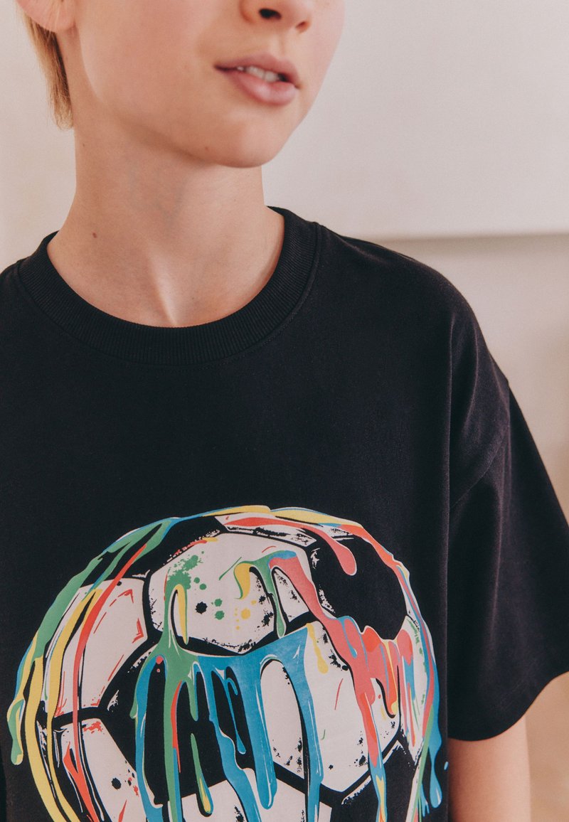 Unge person iført en sort t-shirt med et flerfarvet malet dryppet fodboldmotiv foran.