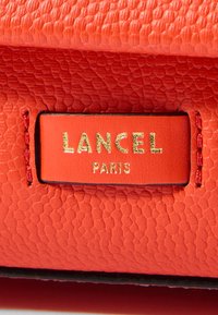 Koralfarvet struktureret lædertaske med et guldfolieret mærke. Mærket lyder "LANCEL PARIS," fastgjort med rød syning på siden.