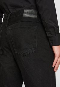 Vaqueros de denim negro con un acabado texturizado, que presentan un bolsillo trasero visible, un parche con el logo en la cintura y detalles de costura sutiles.