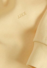 Lys gul bomullsplagg med teksturert overflate og ribbestrikket mansjett, med en diskret pregetetikett "JJXX" på stoffet.